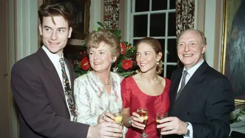 Getty Images Neil and Glenys Kinnock celebrate their 26th wedding anniversary at Temple, London. They are pictured with their children Stephen and Rachel, 26th March 1992. Stephen Kinnock is on the left and in a brown suit with a white shirt and a colourful tie. Next to him is his mother Glenys in a white dress, next to her is her daughter Rachel who has a red dress on. And on the right is Neil Kinnock, who has a dark blue suit on. All of them smile at the camera and are holding a glass of champagne.
