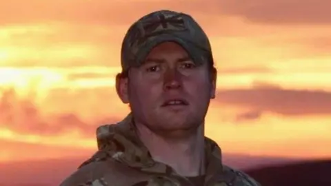 Family handout CPL Sean Roberts, known as Robbo, sees the camera with an orange, yellow and red sunset, which brightens the sky behind him. He is wearing a camouflage jacket and baseball cap, with a green and brown colored association flag above his peak