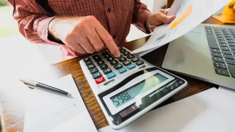 Getty Images Man with bills sits working out his accounts using a calculator 