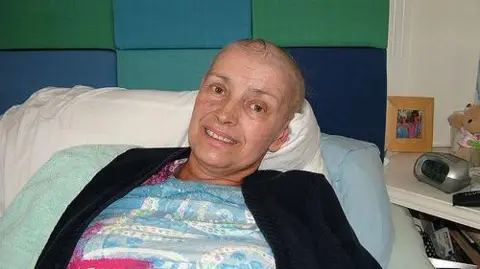 Olwen Kramer Olwen Kramer lies in a hospital bed. She has lost her hair because of cancer treatment. She is wearing a black cardigan with a white, blue and red patterned shirt underneath.  