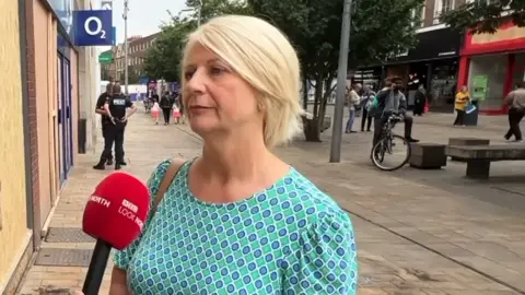 Kathryn Shillito from Hull BID talks to BBC Look North in Jameson Street, Hull. She is wearing a blue and green dress and has blonde hair. There are police officers and boarded-up windows behind her and burn marks on the pavement