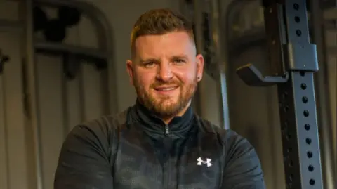 Aidan Smith Man with short hair and a beard looks into the camera wearing a grey, zipped sports top. Behind him are pieces of gym equipment.