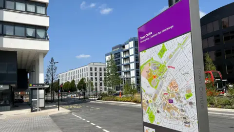 A new sign with a map of Swindon town centre on it. Behind it is a freshly tarmacked road, pavements with plants and trees. Office blocks with large windows surround on a sunny day with blue sky