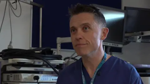A clean cut man is sitting in front of a medical background wearing blue scrubs. He has a light blue NHS lanyard around his neck.