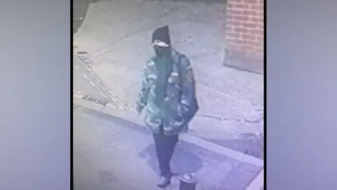 An individual that appears to be a woman wearing a camouflage jacket, head coverings and dark trousers captured on CCTV