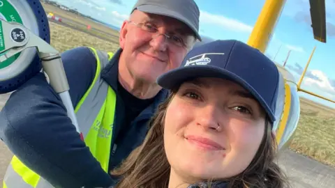 OLIVIA WATSON Olivia and her father on an airstrip with yellow helicopter behind them. They are both wearing blue baseball caps.