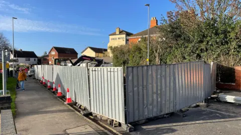 A row of metal fencing and road cones surrounds the roadworks on the A52 Seaholme Road in Mablethopre. Buildings are visible in the background and the fence looks shiny because of the blue sky and sunshine.