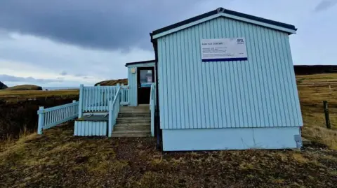 Tammy Hine A blue hut, with steps to it and a ramp. It is clad in panels, with a sign on it. It is surrounded by moss, grass and fields. The sea is in the distance. 