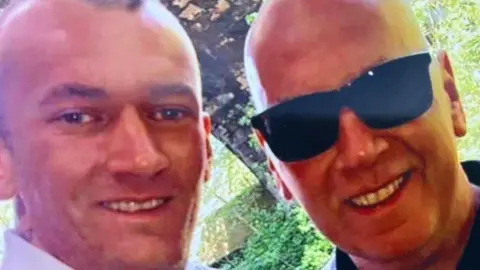 The English family near the selfie style image or two smiling men. One is bald and wears black sunglasses. The other has short and cut hair.