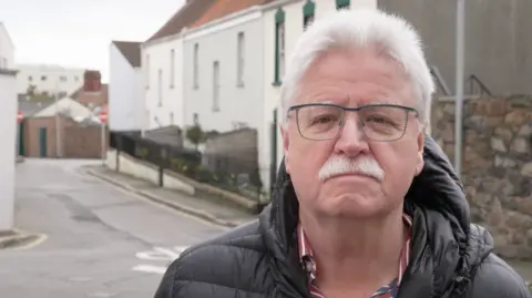 Chris Band: A man in his 60s wearing a black puffer jacket and a blue shirt with a red‑and‑white striped collar is standing in the middle of a quiet street. The background shows a residential area with light‑coloured buildings, a stone wall, and a narrow road curving to the left. The sky appears overcast.
