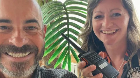 A man and a woman holding a walkie talkie with a beach behind them.