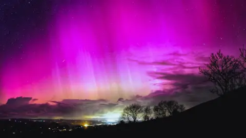 Starry sky illuminated bright pink and moving to peach colour just above low cloud which hangs above the town. The outline of bare trees is clearly visible in the light on the hills to the right.