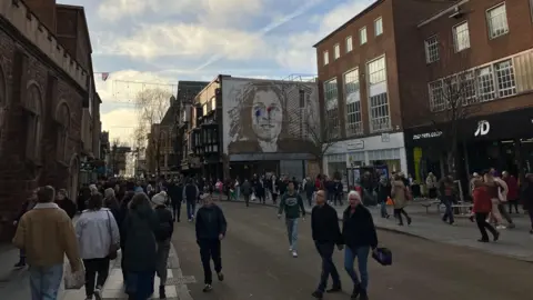 A general view of a busy street in Exeter. Crowds of people are walking along the pavements. Some people are crossing the road. Tall buildings line either side of the street. A mural of a woman's face is painted on the side of one building. 