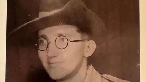 Contributed A black and white image of a young man wearing a cowboy-style hat and glasses. He is looking away from the camera and slightly smiling. 