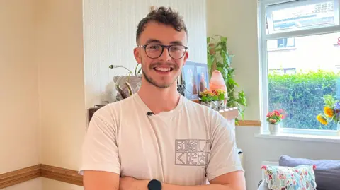 Conor Finnerty, wearing glasses and a white T shirt,  smiling at the camera in a living room