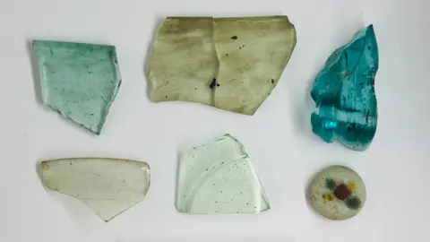 AC Archaeology and Heritage Six shards of glass. All have various levels of translucence. The top left is light green in colour and you can see through it. The top middle is clear but almost looks smeared with a dirty brown material. The top right is the most striking being the thickest shard and a ice cold blue. The bottom left and middle are the translucent with specs of black on them. The bottom right is the least translucent with a number of what look like beads in the glass. There's a red one in the middle two yellow ones North East and South West and two greens ones South East and North West. 