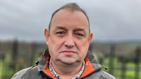 Declan Norris has short brown hair.
He is wearing a checked shirt and a green-and-orange hooded rain jacket.
The background has been blurred.