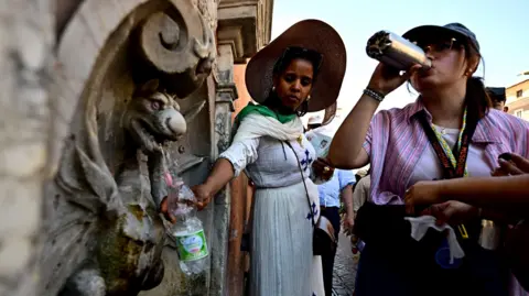 AFP People wearing sunhats fill water bottles and drink them at a water fountain in Vatican City on 28 June, against a blue sky.
