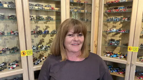 A woman standing indoors in front of wooden display cabinets with glass shelves. The cabinets are filled with numerous small model racing cars arranged neatly in rows. Yellow signs reading “PLEASE DO NOT TOUCH” are visible on the display cases, indicating a collection or exhibition setting.