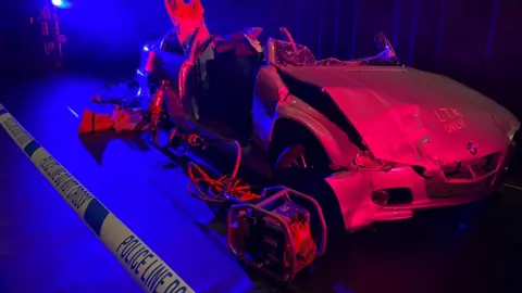 Guernsey Ambulance and Rescue A crashed car behind police line "do not cross" tape. The car is part of the presentation exhibition. It is mangled and with red light being shone on it. The roof is missing and one of the doors has been taken off 