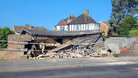 Karen Vilday Crumbled bricks on the floor on the same section of wall from 1 June last year. Police tape is stretching across between the two sections of wall which are still standing. The garden shed is also damaged with one of the walls caved in. Behind there is a large white house which is next to Mrs Vilday's house. The skies are blue and clear 