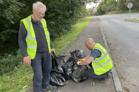 Two volunteers in yellow vests collect black bags of rubbish - one is standing looking at the camera, the other is crouching down to tie the top of one of the refuse sacks.