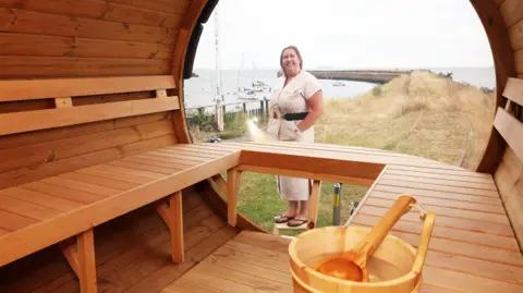 The image shows the inside of a wooden style sauna, Kathryn Donavan, the owner of the sauna is stood just outside. She's wearing a beige dress with a black belt and a pair of black flip flops. She has short blonde hair.