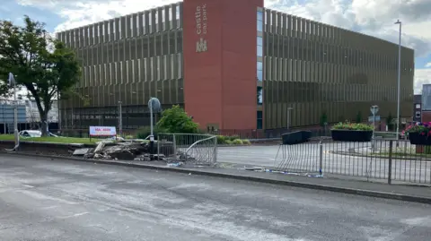 A street scene shows a damaged brick wall and police tape, suggesting a recent crash. A tree and the "Castle Car Park" can be seen in the background. 