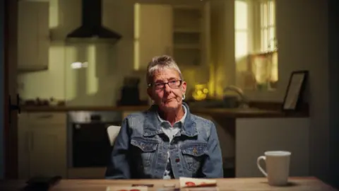 Yeti Television/BBC Cymru Wales Photograph of Tarnia Holme sat in her kitchen. She is in her 60s/70s and has short silver hair and wears black rectangle glasses. She wears a denim jacket and looks directly at the camera, pursing her lips. A cup of tea can be seen on the table in front of her. 