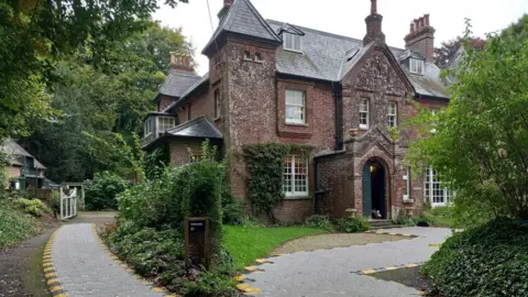 Max Gate is a large stately dark brown brick manor house, with a grey tiled roof. It is surrounded by greenery, with trees, hedging and grass framing the property and a safety pathway has been installed to lead people to two different entrances. The pathway is lined with yellow and black paint.