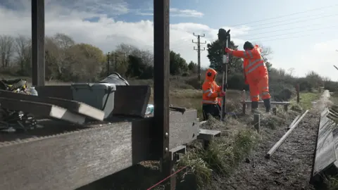 BBC / Kevin Church Two men in orange high-vis outfits and protective headsets are working on the path. They are standing with one of them holding a pile and the other one drilling the pile into the ground. In the foreground, a wooden walkway is seen and there are the men's tools on it. 