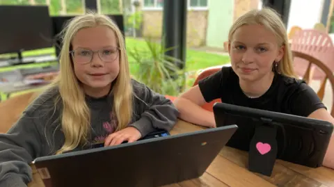 Rosa est une fillette de 8 ans aux longs cheveux et lunettes blonds. Elle porte un pull gris et rit. À sa droite, sa sœur de 14 ans Matilda, avec de longs cheveux blonds, portant un cycle d'oreille en argent et un t-shirt noir. Ils sont tous les deux assis à la table, ont des appareils devant eux et sourient.