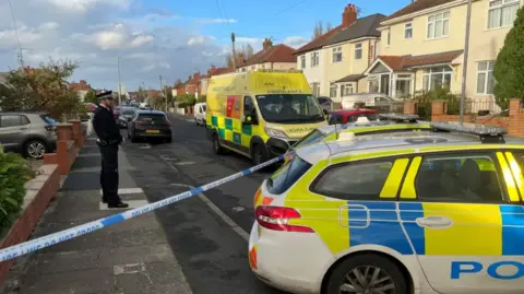 A male police officer in black uniform stands guard on a residential road, a line of police tape and a police patrol care is in the foreground and a yellow ambulance is parked a few yards away from the officer. 