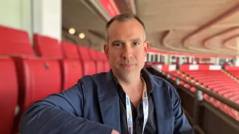 A middle-aged white man wearing a blue jacket and black shirt, sitting with his arm resting on the back of a seat inside a football stadium