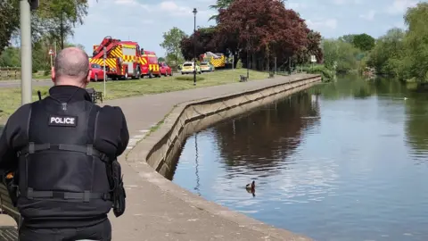 PURPLEVISION A police officer guards the River Nene at the embankment in Wellingborough. Rescue vehicles can be seen in the background.