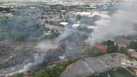 Drone footage of smoke rising from a grassland area. A large car park, playing field and buildings including houses can be seen beyond.