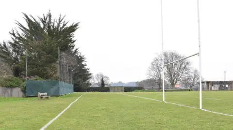 Crown Prosecution Service A rugby post on one side of a recreation ground, bordered by trees and houses