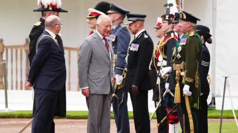 PA Media King Charles III wearing a grey suit and white shirt walks with the Lord Provost of Edinburgh Robert Aldridge, who wears glasses and a dark-coloured suit. The pair are greeted by uniformed figures.