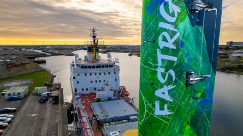 Smart Green Shipping A close-up drone view of the tall rectangular green and blue sail fitted to the deck of the cargo ship. It has the word 'FASTRIG' written vertically on it The ship's funnel and bridge can be seen in the background.