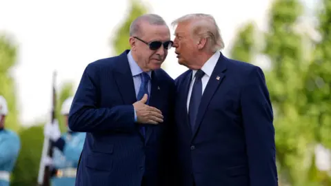Donald Trump, dressed in a navy suit and tie, talks into the ear of Turkish President Recep Tayyip Erdogan who is also wearing a blue suit and tie, as well as sunglasses.