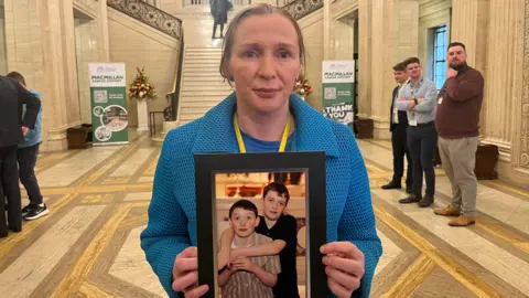 Colleen McConville holds a photograph of her two sons, Cian and Cillian, as she looks at the camera with a neutral expression on her face. She is wearing a patterned blue coat. Her light hair is tucked back behind her ears.
