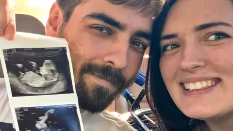 A man with short dark brown hair and a beard sits next to a woman with shoulder length brown hair on a hospital bed.  He is holding up baby scan images to show the camera.