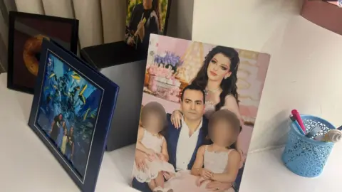 A family photo showing Khodr Taher Bin Ali and his wife with his two children, their faces blurred. The photo frame is on another table, with other photos nearby