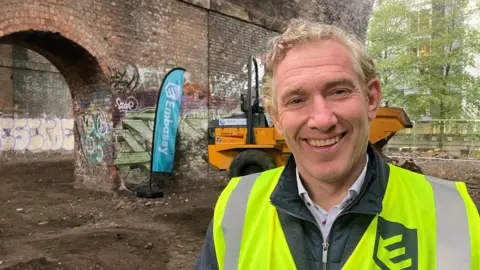 A smiling James Whittaker from Peel Waters stands underneath a series of railway arches in the Castlefield area of central Manchester. He is wearing a yellow hi-vis jacket.