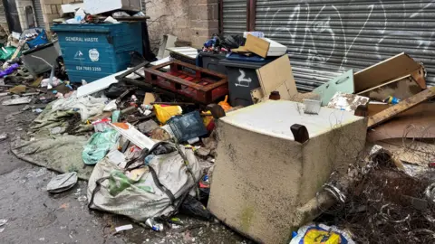 A large pile of rubbish which is surrounding a rectangle shaped bin. There is an upturned arm chair and other furniture. A plastic Lidl bag, other bags, materials and other waste. 