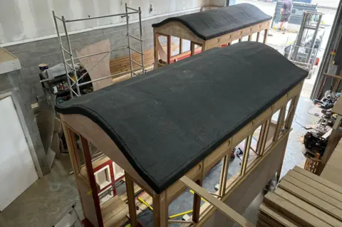 David McKay The two carriages placed diagonally from one another in the warehouse. The wood making up the frame is still light brown and the roof is dark. The windows all around the structure are bare. There is construction equipment surrounding them.