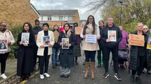BBC A number of parents and carers stand in the road holding signs and pictures of children who have been supported by Richard House. Some signs read "Save Richard House"