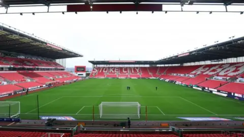 The view onto the pitch from the stands of a large football ground. The seats are red, with Stoke City picked out in white on the opposite side stand