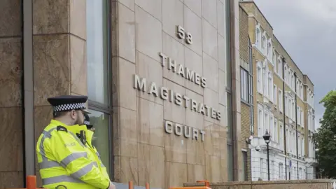 Two police officer in a high-visibility yellow jackets stand in the foreground, at the the entrance of Thames Magistrates' Court. The building's stone façade features the court's name in large, metallic lettering below the number 58. To the right, a traditional brick building with white window frames and a black lamp post lines the street.