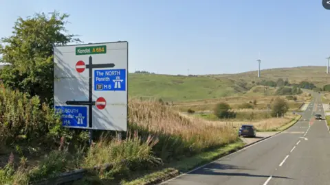 A general view of the approach to Junction 37 of the M6 from the A684. The single carriage road is in a rural setting, surrounded by fields and hills. A sign at the side of the road indicates the junctions.